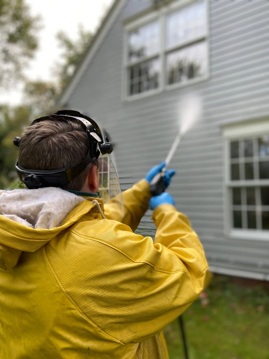 Photo by Caitlin Whealy Person cleaning house siding with a pressure washer, wearing protective gear in a yellow raincoat.